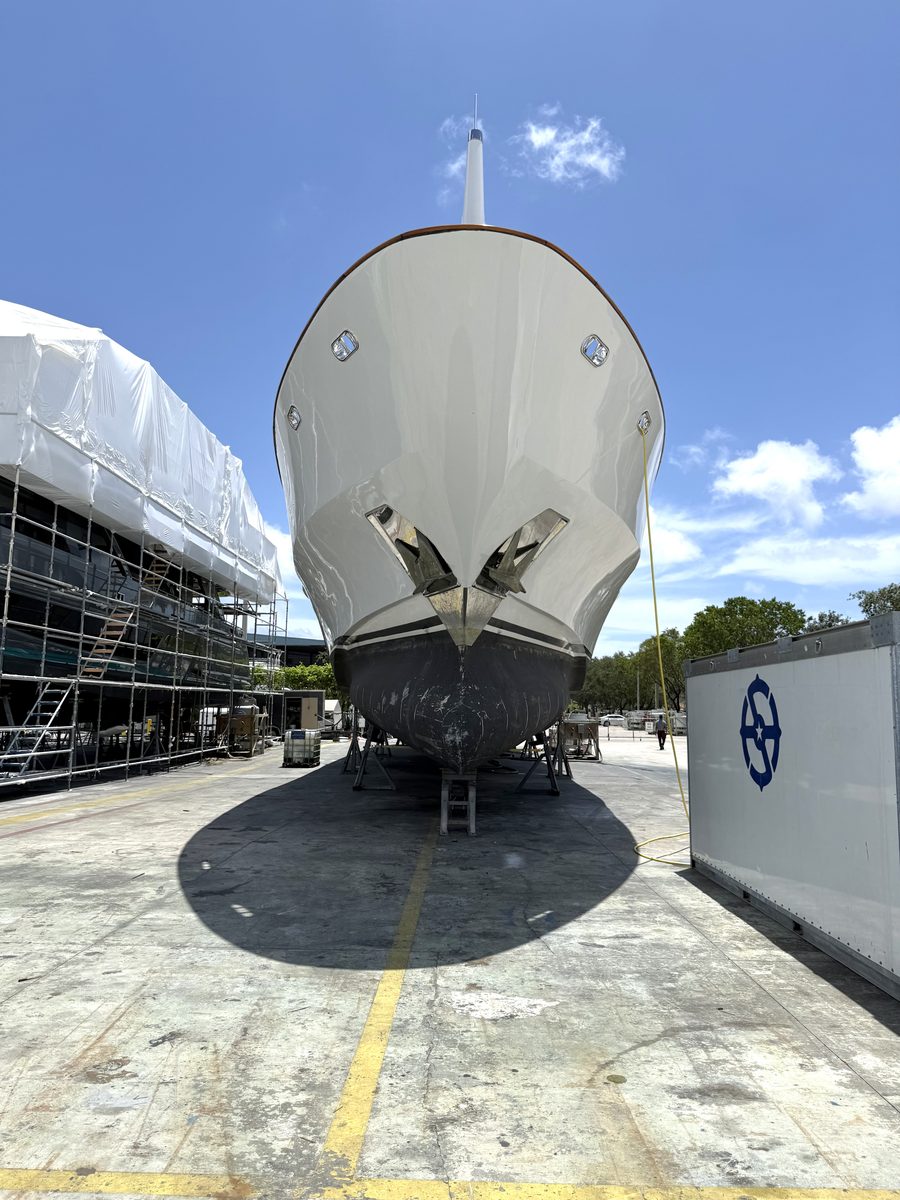 The bow of a white motor yacht towering overhead on jack stands in a working boatyard under a blue sky.
