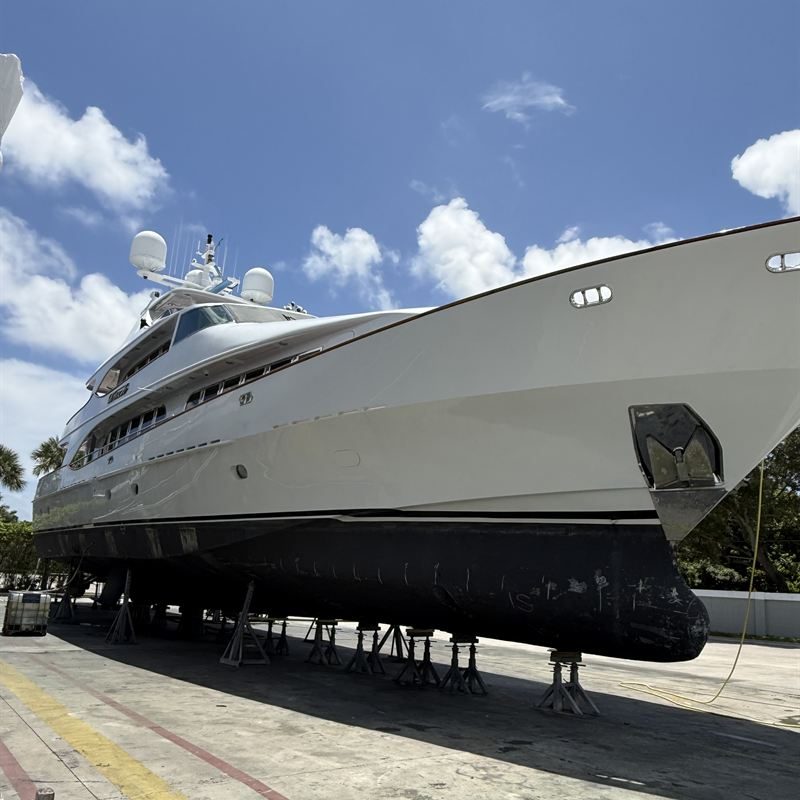 A large white motor yacht standing on jack stands in a boatyard, its underwater hull fully exposed.