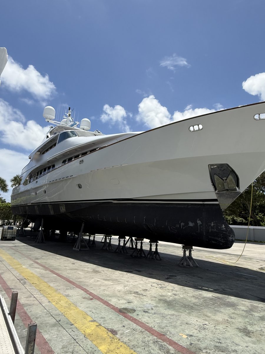 A large motor yacht standing on blocks in a boatyard, the vessel's entire underwater hull exposed, photographed from beside the bow.