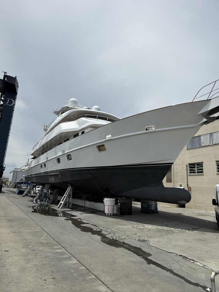 A large white motor yacht hauled out in a working boatyard, the vessel's dark underwater hull fully exposed beside a travel lift.