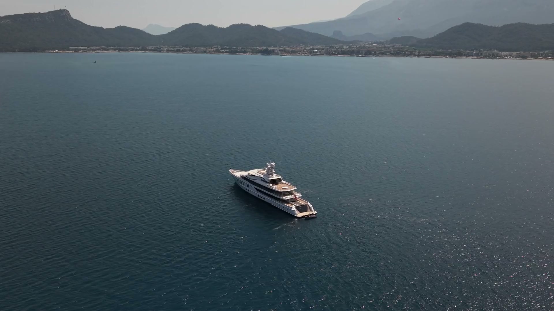 A superyacht under way on open blue water, a mountainous coastline and hillside town behind the vessel.
