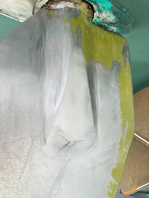 A rudder blade with antifouling worn back to bare metal, and green corrosion bloom at the stock and bearing above it.