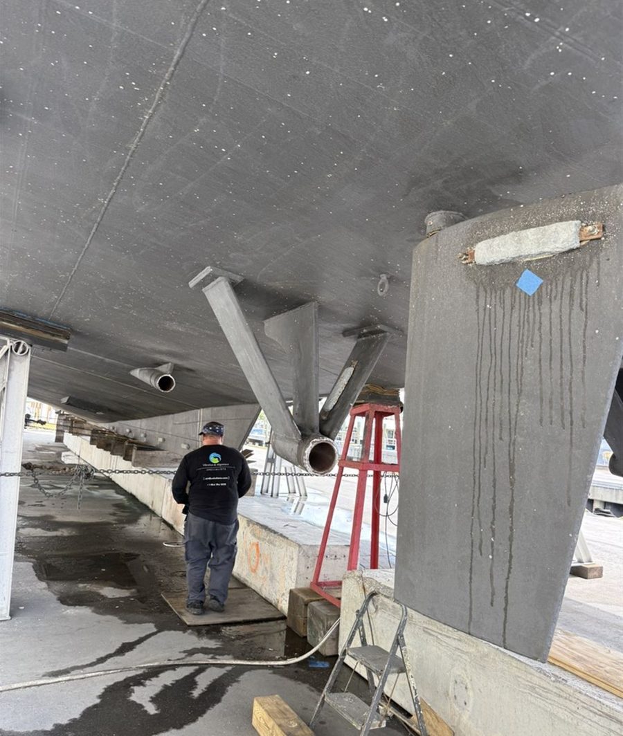 The underside of a hauled vessel showing shaft struts, through-hulls and a sacrificial anode on the rudder, with a person walking beneath the hull.